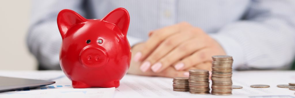 A red piggy bank sits in the foreground with stacks of coins to the right, while a person's hands rest on a table in the background.