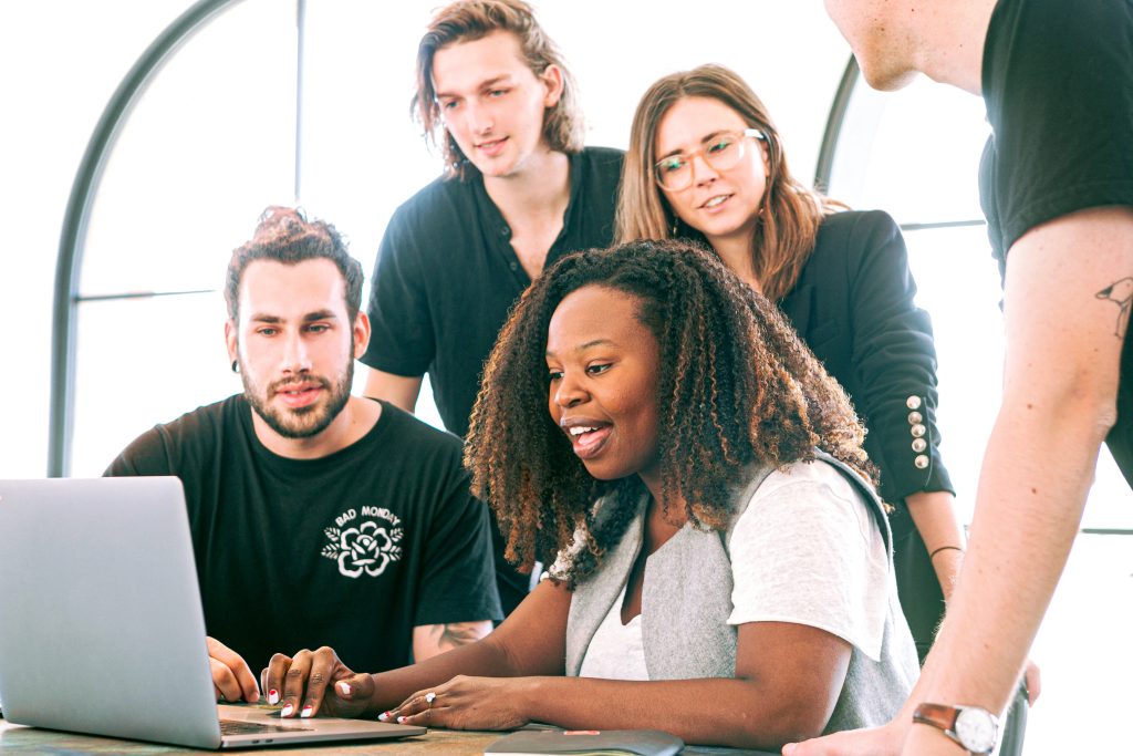 A group of young adults gathered around a laptop, engaging enthusiastically in a group discussion inside a brightly lit room with a large window.