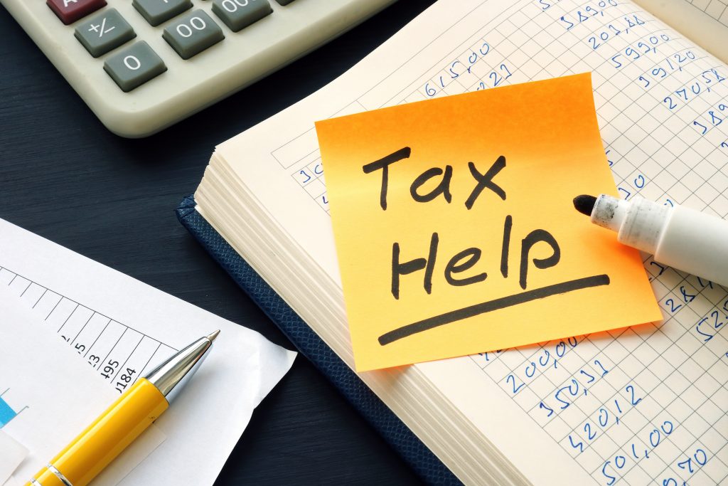 A sticky note with "tax help" written on it, placed on a desk with financial documents, a calculator, a pen, and a highlighter.