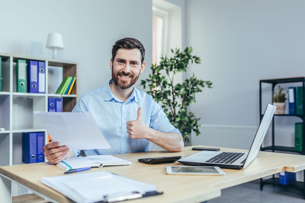 A smiling man with a beard in a casual shirt sits at a desk, giving a thumbs up while holding documents. a laptop and smartphone are nearby, in a neat office with bookshelves and plants.