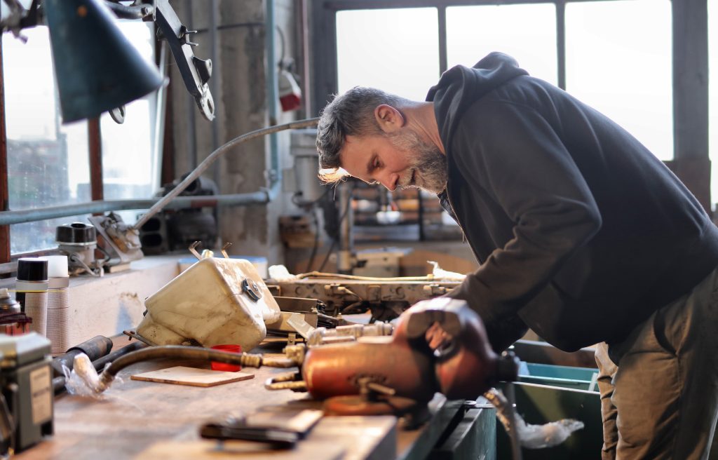 A man with a beard works intently in a workshop, sanding a curved wooden object while surrounded by various tools. the setting is warmly lit, suggesting a focused yet creative atmosphere.