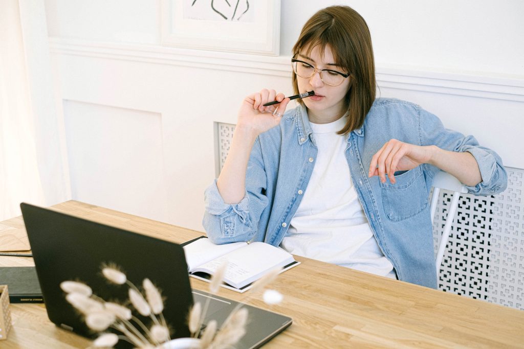 A woman in glasses, wearing a denim jacket over a white shirt, sitting at a desk with a laptop and notebook, pensively holding a pen near her mouth.