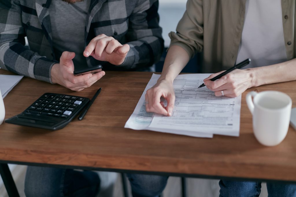 Two people seated at a desk, reviewing documents and using calculators. one person holds a smartphone, and a coffee cup is visible on the desk.