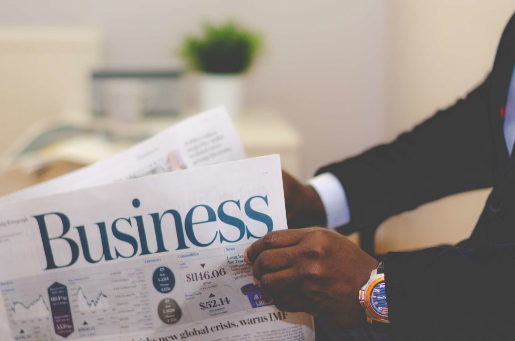 A person in a suit reading a business newspaper, with focus on their hands holding the paper and a visible wristwatch. the office setting is softly blurred in the background.