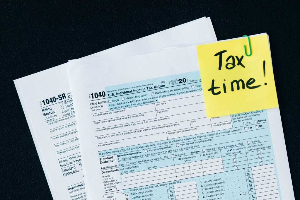 Several U.S. tax forms (including 1040 and 1040-sr) are spread on a dark surface, with a yellow sticky note that says "tax time!" in green marker.