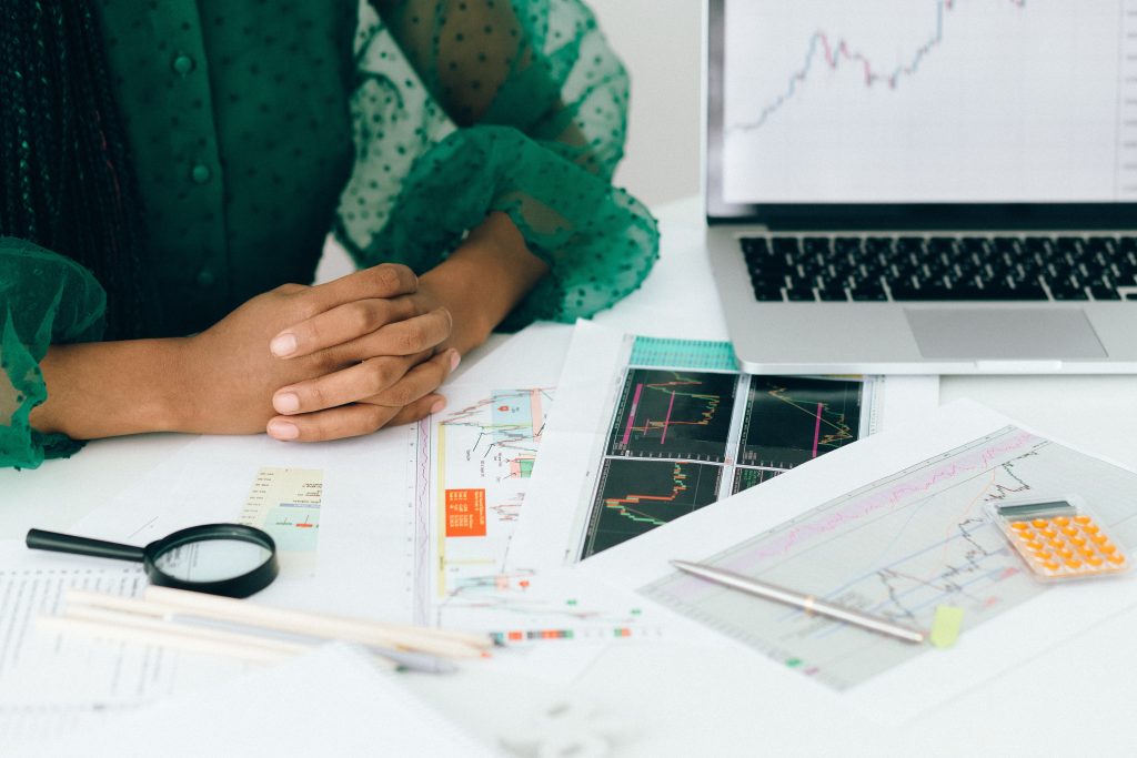 A person in a green blouse sitting at a desk with a laptop displaying graphs, surrounded by financial charts, a calculator, and a magnifying glass.