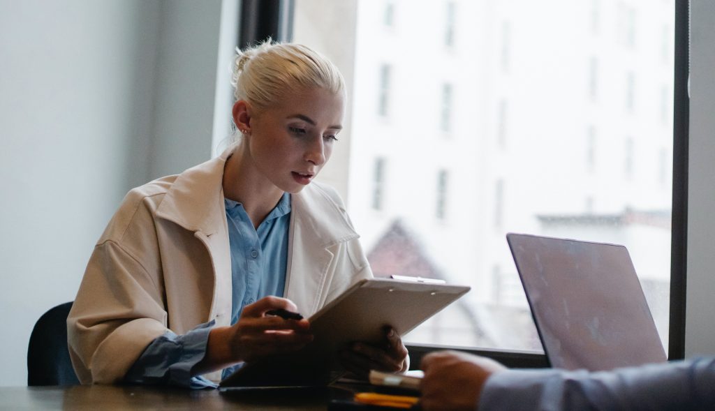 A focused woman with blonde hair, wearing a light coat, sits at a desk holding a stylus and digital tablet, possibly in a business meeting, with another person partly visible across from her.