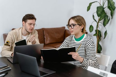 Two colleagues discussing work while looking at a laptop and a notebook in a modern office setting with a potted plant in the background.