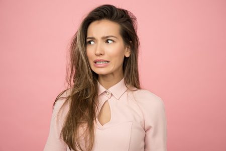 A young woman with long brown hair and braces wearing a pink blouse looks thoughtfully to her right, a concerned or worried expression on her face. All set against against a pink background.
