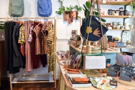 A cosy boutique shop interior featuring a clothing rack with colorful garments, shelves with various bags, and a display of small items like wallets and decorative plants.